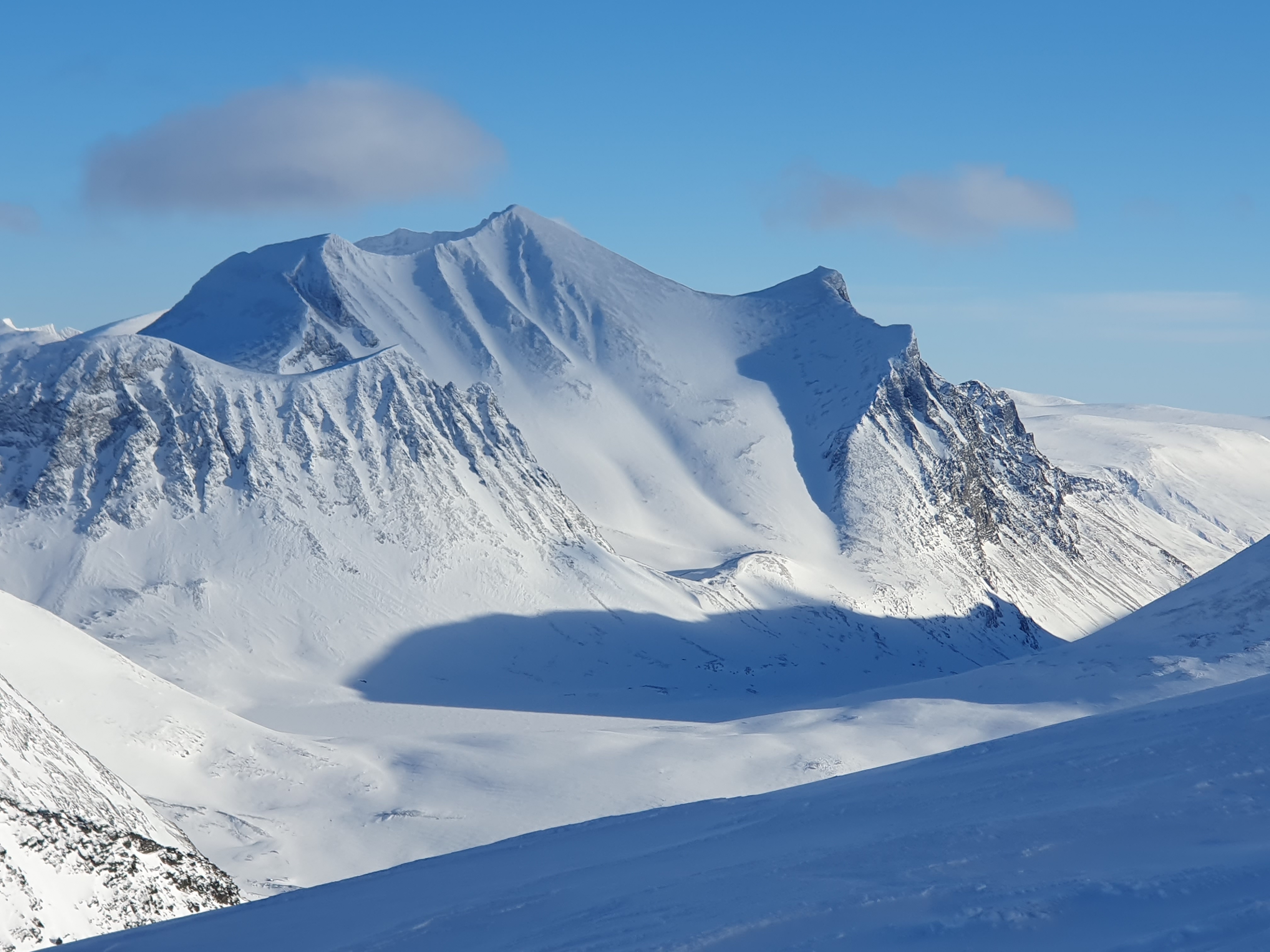 Utsikt från ett fjäll över en snöklädd dalgång. Ena berget ger skygga i dalen. Blå himmel bakom.