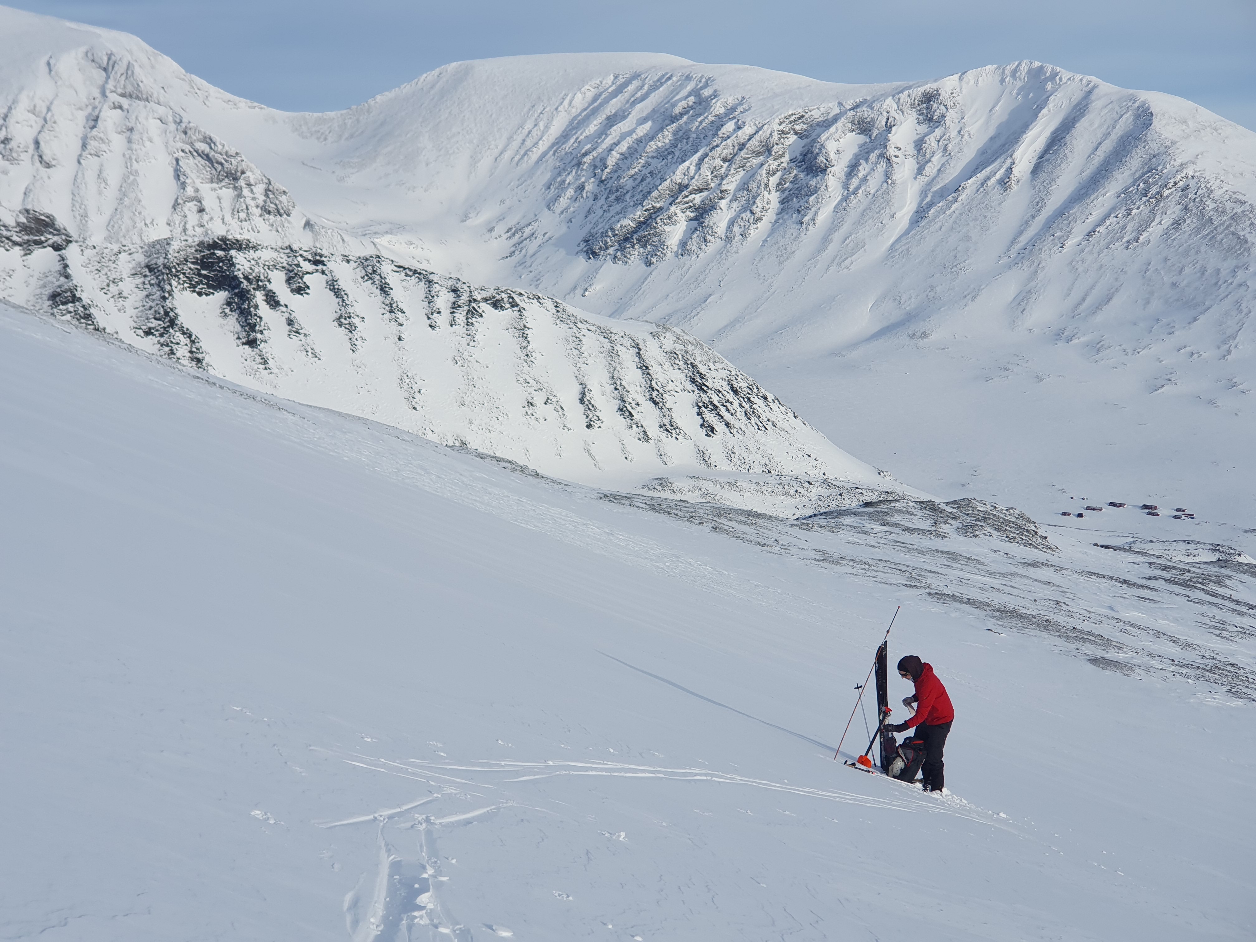 Snowboardåkare med röd jacka och svarta byxor står mitt i backen och fixar med sin utrustning. Man ser fler snöklädda fjäll i bakgrunden och blå himmel.