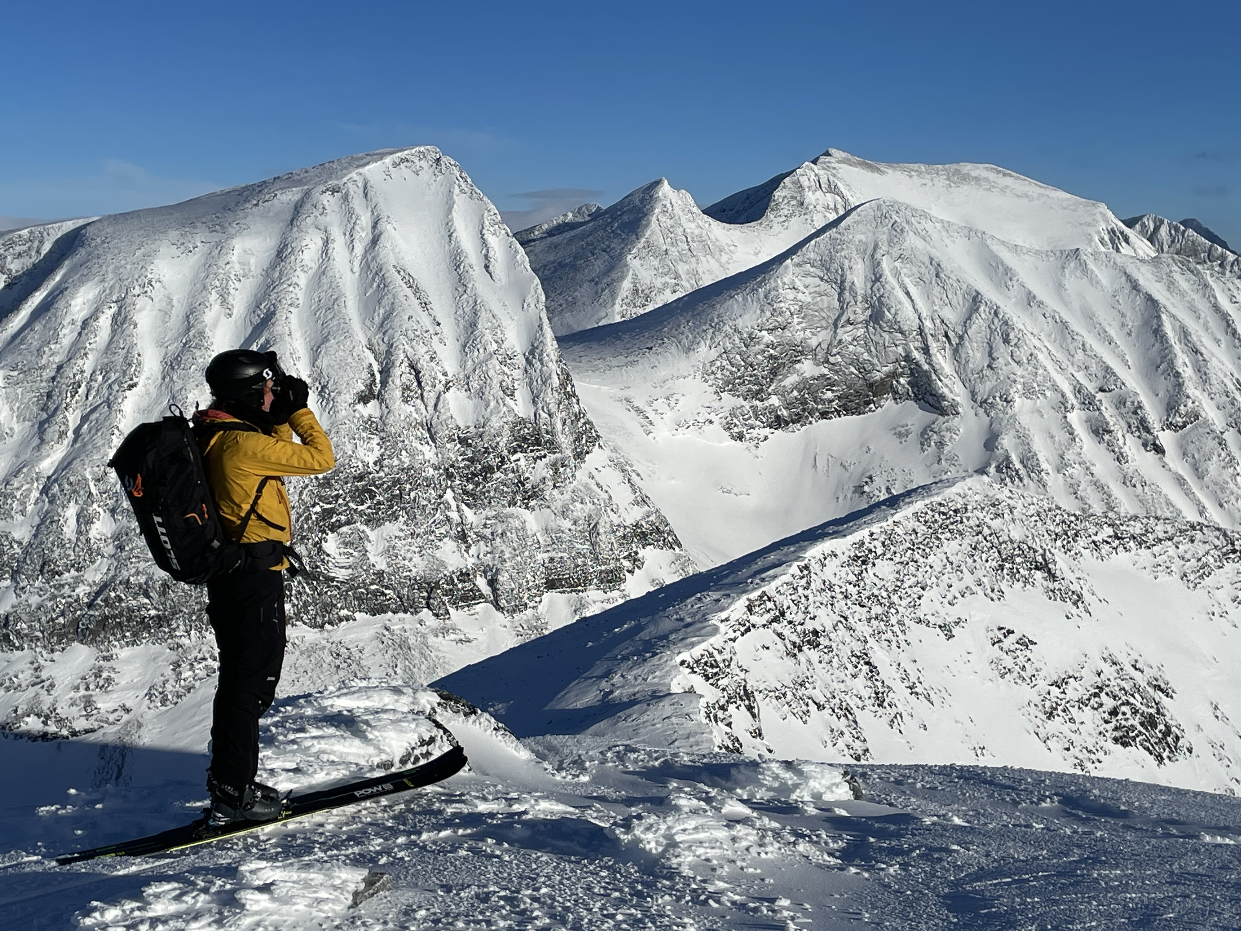 Kvinna i gul jacka med rycksäck och skidor på fötterna blickar ut över snöklädda fjäll.